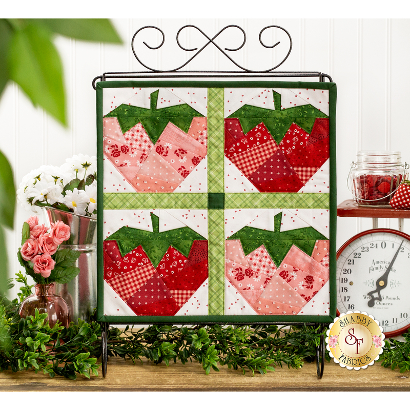 The completed June wall hanging, a pink, red, and green collage of four juicy strawberries, hung on a craft scroll and staged on top a rustic wooden shelf with coordinating decor and flowers.
