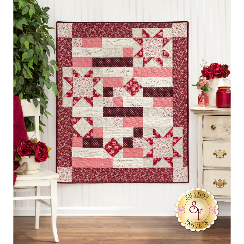The completed Comfort of Psalms quilt in Blushing Blooms, a collection of red, pink, and cream floral fabrics. The quilt is hung on a white paneled wall and staged with coordinating furniture and decor, including red and pink roses.