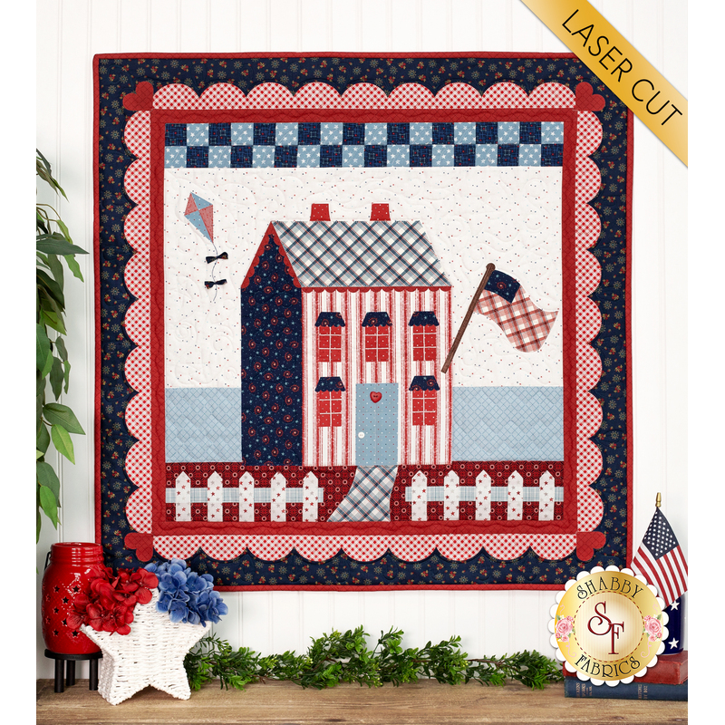 The completed Little Garden House in America wall hanging in a patriotic palette of red, white, and blue, hung on a white paneled wall and staged with coordinating decor and leafy plants.