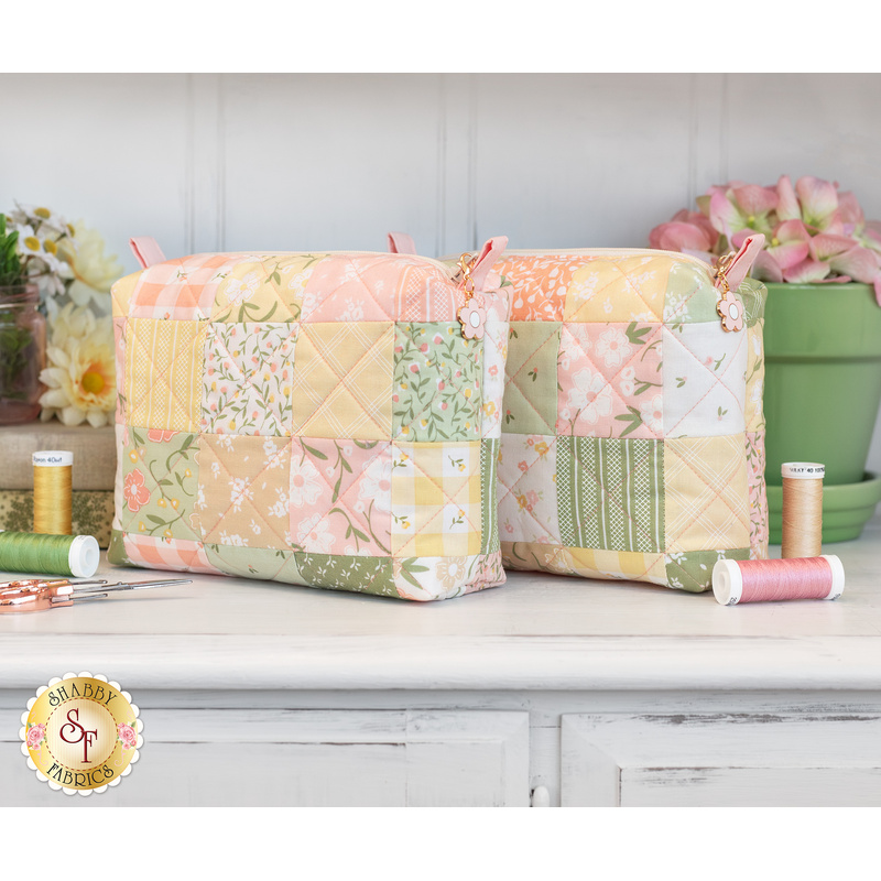 The two completed mini charm bags in pastel shades of green, yellow, pink, and white. The bags are staged on a light wooden counter with coordinating spools of thread and flowers.