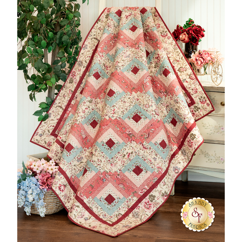 A cream, light blue, and baby pink colored quilt with log cabin blocks draped over a dresser next to a green shrub