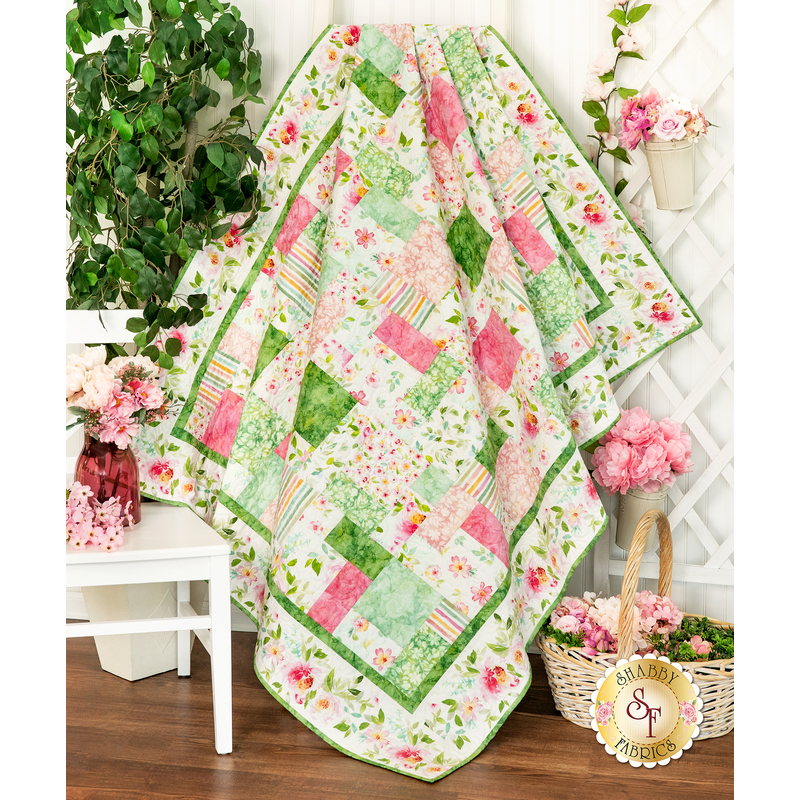 Photo of a finished pink, white, and green floral quilt draped over a white ladder in front of a white wall, houseplants, pink flowers, and a white chair and trellis.