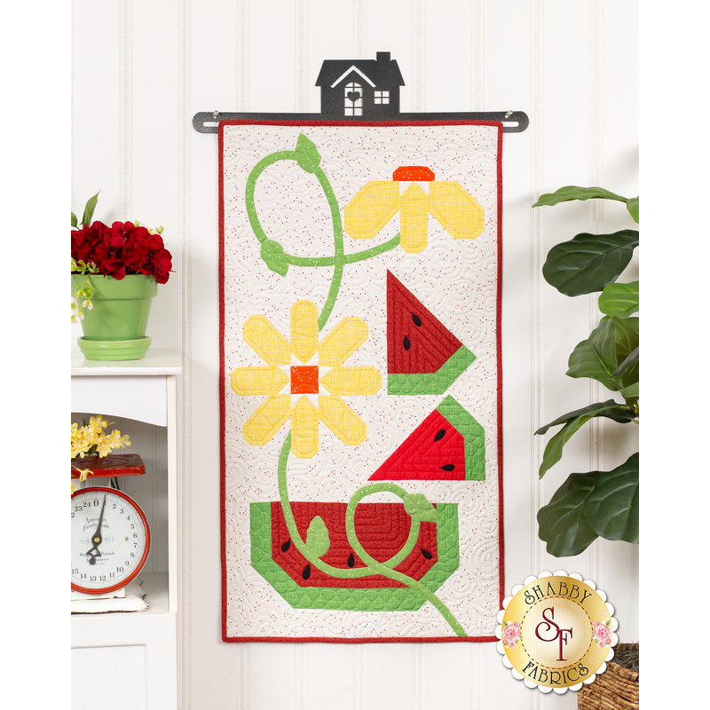 The completed door banner for August, a twisting tableau of yellow flowers and watermelon slices, hung on a white paneled wall and staged with coordinating decor and a leafy houseplant.