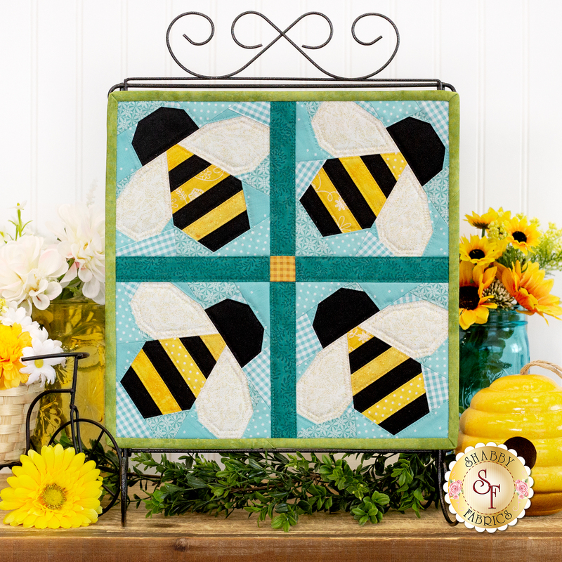 The completed Foundation Paper Piecing August project, colored in bright aqua and yellow fabrics, staged on a rustic wood set of drawers with coordinating flowers and decor.