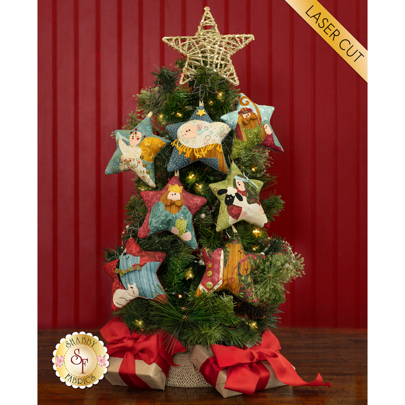 The completed Star of Wonder Nativity ornaments staged on a small Christmas tree against a red paneled wall. Small kraft paper presents with red ribbon sit under the tree and a yellow banner in the upper right hand corner reads 