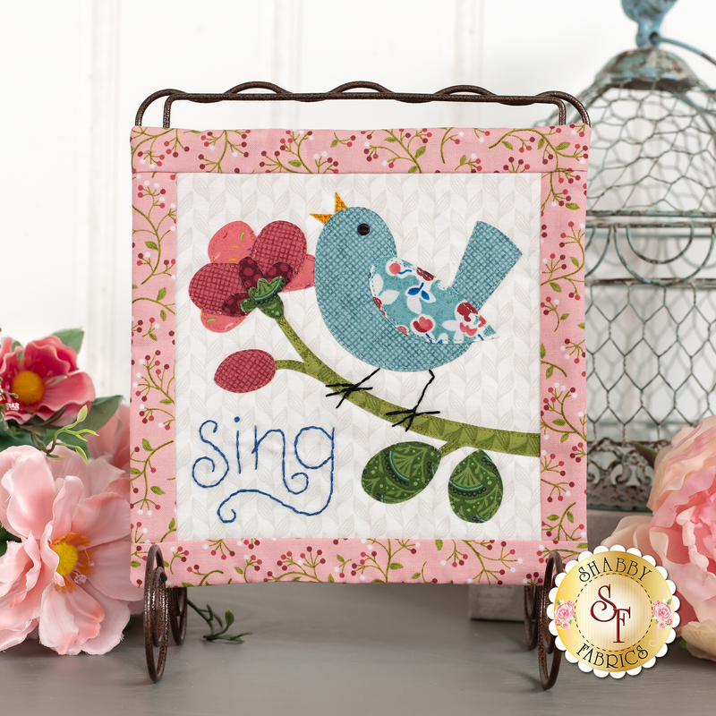 The completed Blossom Trail - Sing Mini Quilt, hung on a craft display and staged with coordinating flowers and decor.