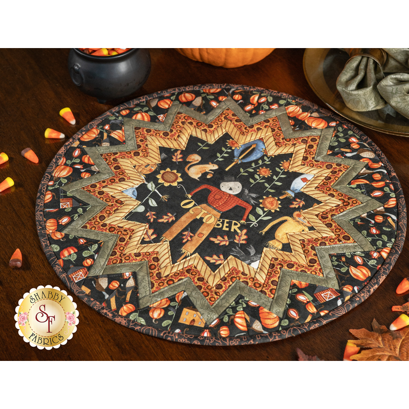 A closer shot of the completed Point of View Folded Star Table Topper in warm fall colors from the Bittersweet Farm collection. The Topper is staged on a brown table with fall leaves, candy corn, and sage green cloth napkins on black and gold plates. The bottom of a pumpkin and small plastic cauldron filled with candy corn peek down from the top of the shot.