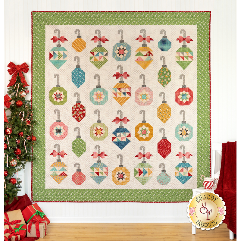 The completed Decorating The Tree quilt, a colorful quilt featuring ornament designs, displayed on a wall beside a decorated Christmas tree.