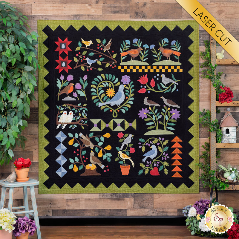 The completed Aviary quilt, hung on a rustic wood panel wall and staged with coordinating foliage, flowers, and decor. A yellow banner in the upper right hand corner reads 