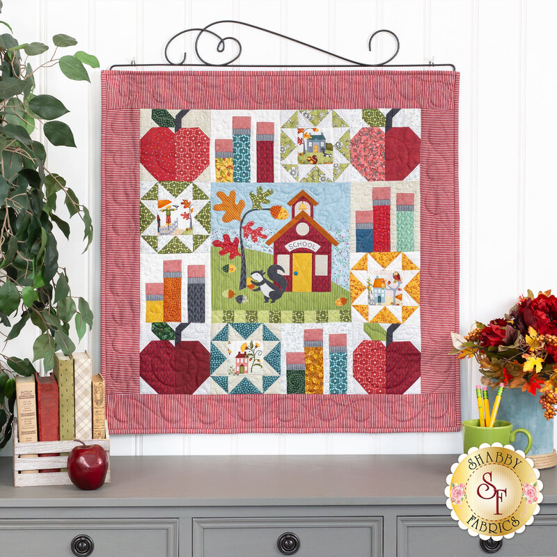 The completed Schoolhouse Haven wall hanging, hung on a white paneled wall and staged with coordinating flowers and school decor.