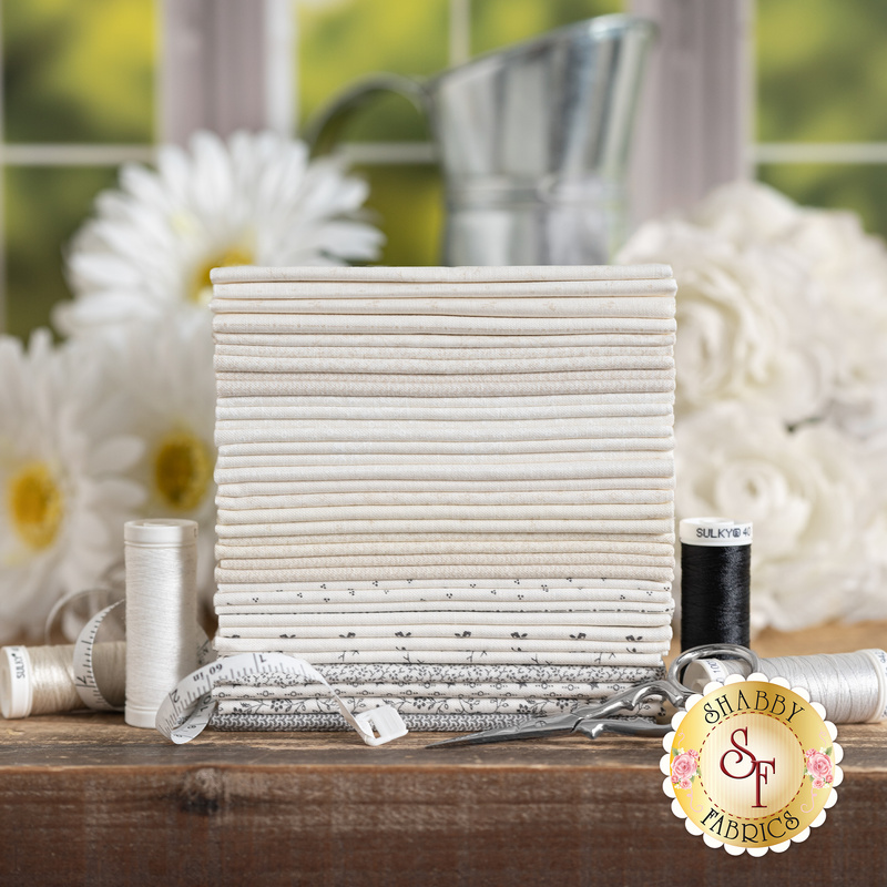 Photography of a stack of neutral fabric rolls with spools of thread and a measuring tape on a wooden table, surrounded by flowers.