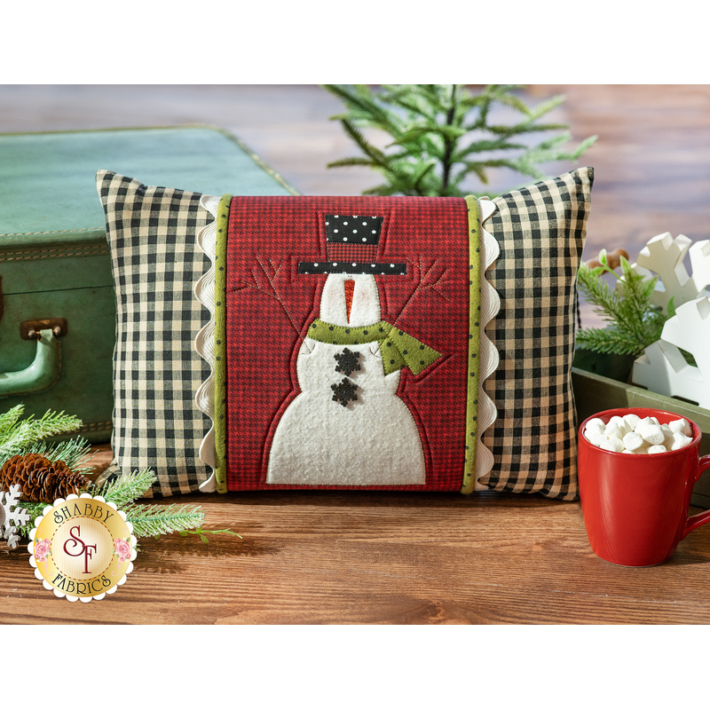 The finished January Pillow Wrap & cover, staged on a rustic wood floor beside a vintage suitcase and a red mug of hot chocolate.