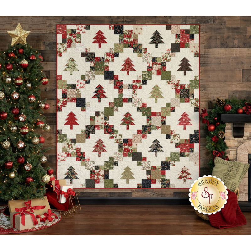 A winter themed cream quilt with evergreen trees and green, red, and brown fabric blocks making a lattice pattern, staged on a wood panel wall with a decorated Christmas tree.