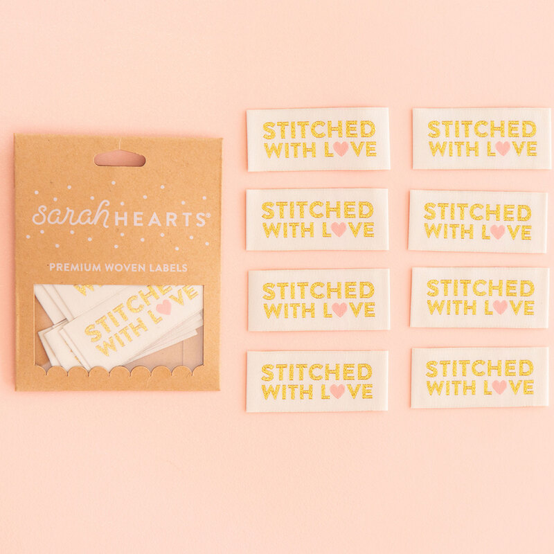 A package of Stitched With Love Sew In Labels next to 8 individual labels on a pink background to show scale and detail.