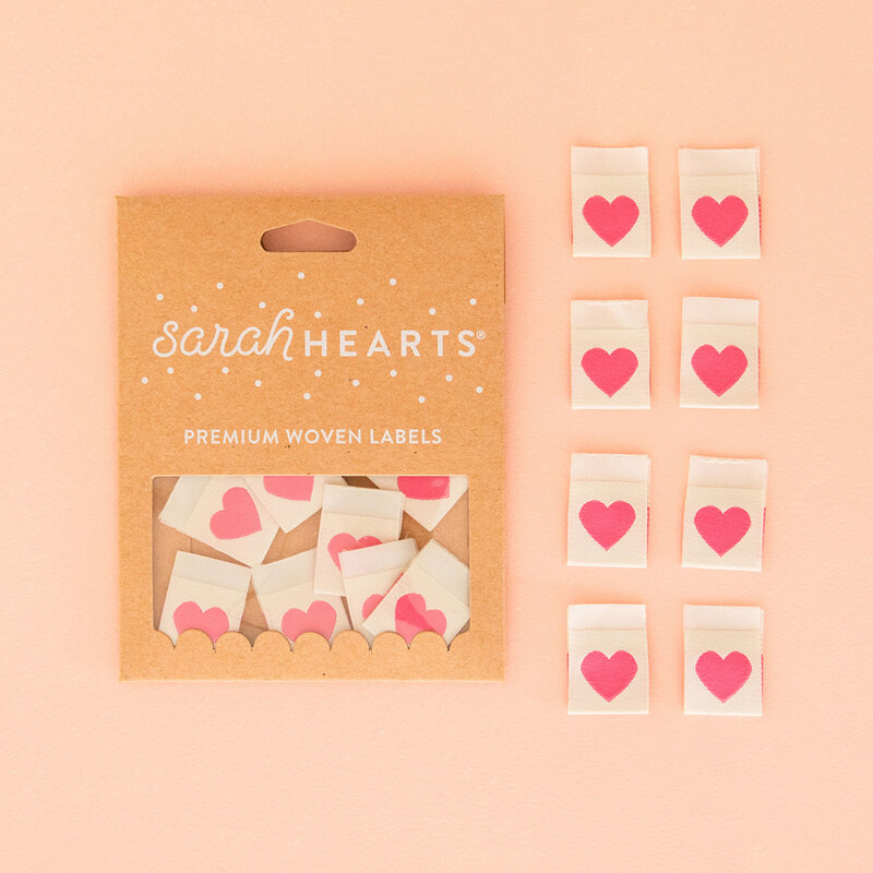 A package of heart Sew In Labels next to 8 individual labels on a pink background to show scale and detail.