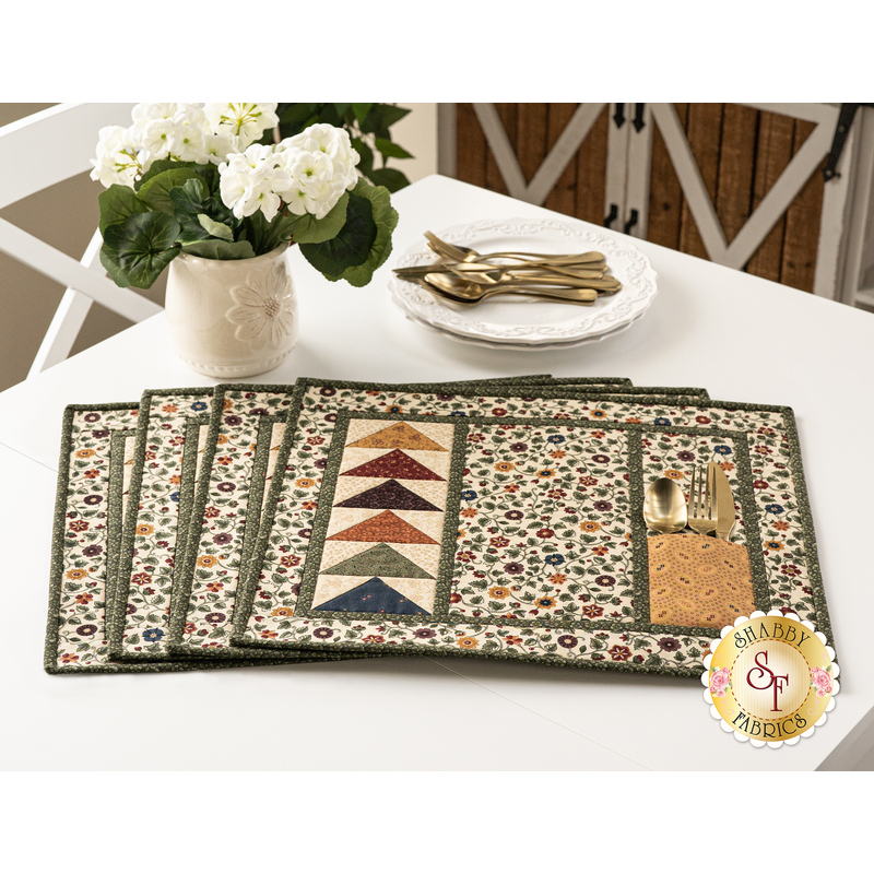 The completed Chickadee Landing placemats, slightly fanned out, staged on a white table with elegant white flowers and tableware.