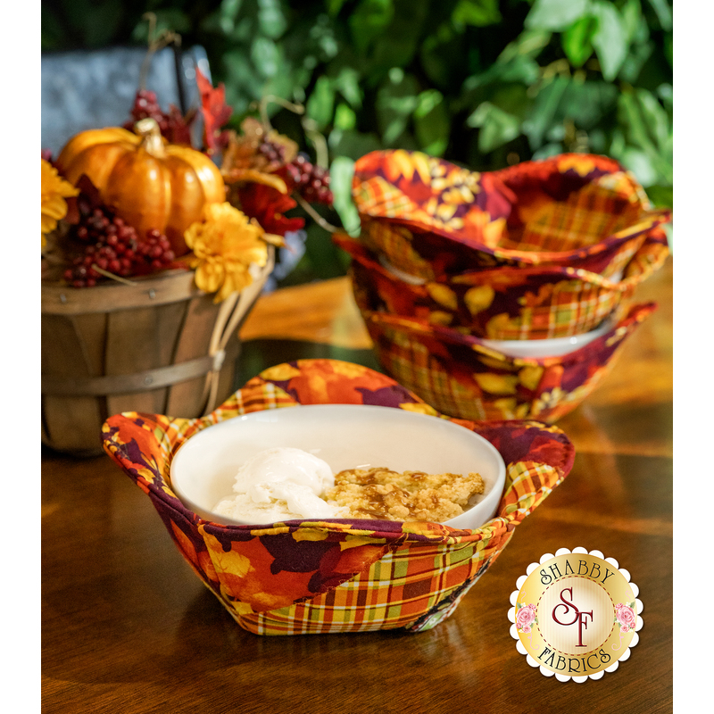 A vibrant fabric bowl cozy filled with dessert, surrounded by decorative fall items and a stack of three other matching bowl cozies.