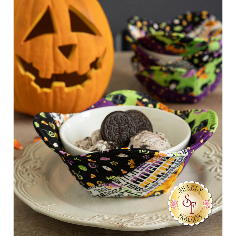 A decorative fabric bowl with ice cream and cookies, set against a backdrop of coordinating decor like a carved pumpkin, candy corn, and three other bowl cozies.