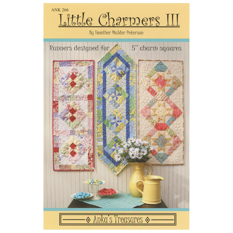 The front of the Little Charmers III pattern showing the finished table runners on a cream paneled background with coordinating colorful decor on a dark brown wooden table in front of the runners.