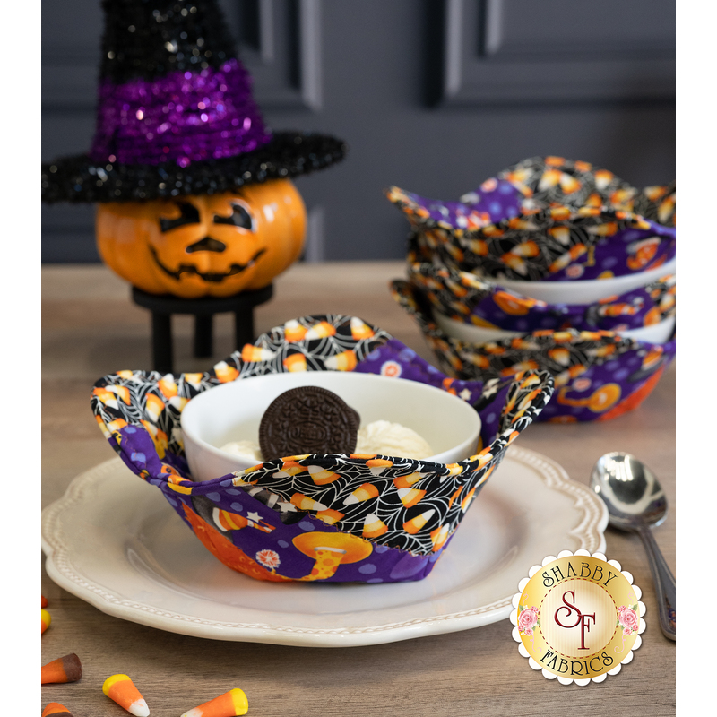 A vibrant fabric bowl cozy filled with dessert, surrounded by decorative Halloween items and a stack of three other matching bowl cozies.