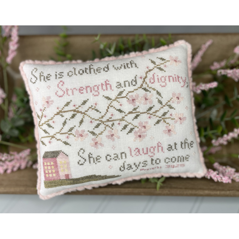 A decorative small pillow featuring a house, vines and flowers including a stitched passage from Proverbs 31:25 