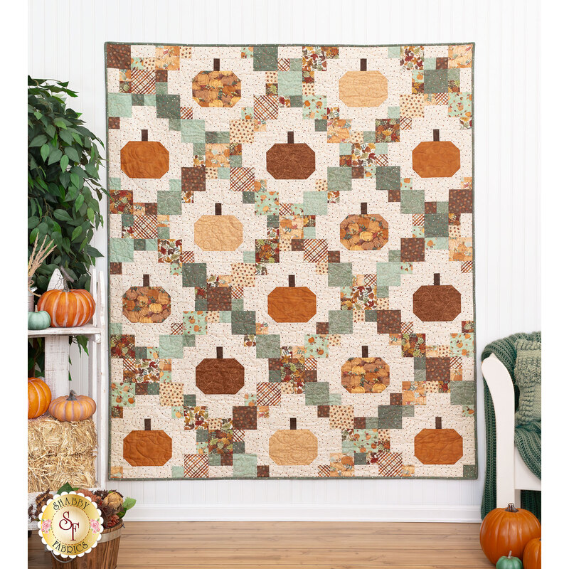 The completed Pumpkin Patches Quilt, colored in elegant and muted colors like cream, burnt orange, and eucalyptus, hung on a white paneled wall and staged with pumpkins and coordinating decor.