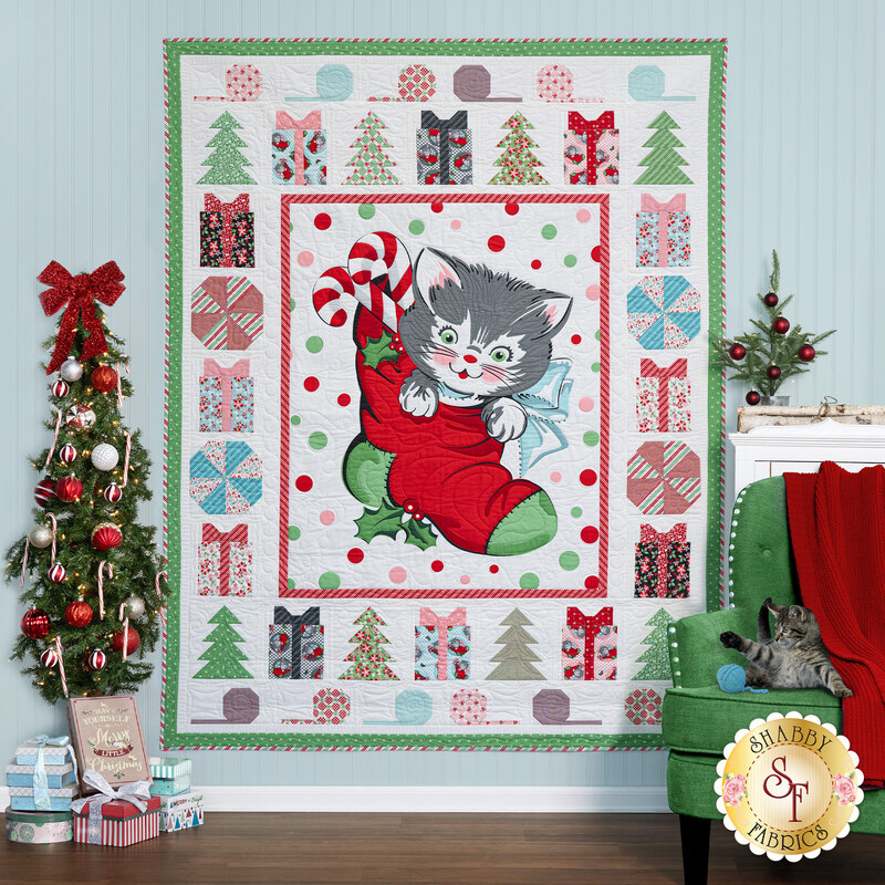 The completed Meowy Christmas quilt, colored in bright green, red, and blue fabrics, hung on a paneled wood paneled wall and staged with coordinating Christmas decor.