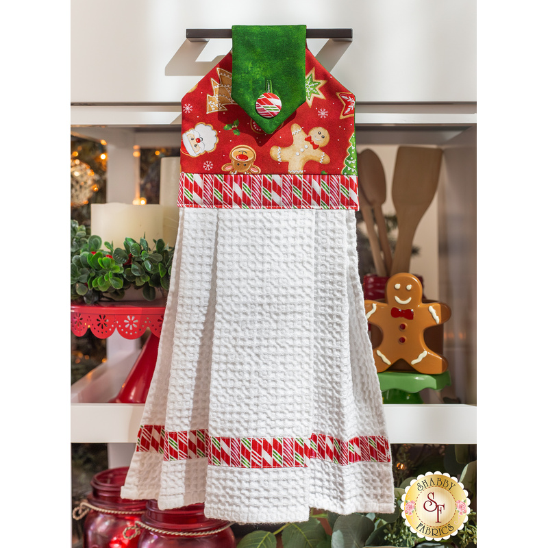 The completed hanging towel in red, hung from a kitchen drawer and staged with coordinating decor like a candle, cooking utensils, and a gingerbread man.