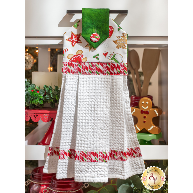 The completed hanging towel in white, hung from a kitchen drawer and staged with coordinating decor like a candle, cooking utensils, and a gingerbread man.