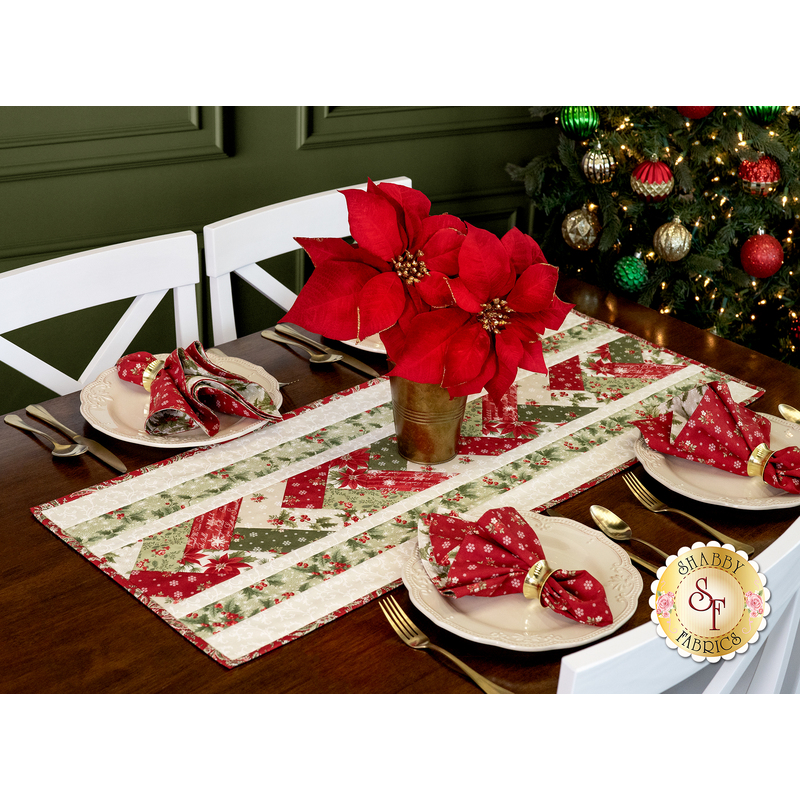 The completed Venice Table Runner, colored in red, cream, and green fabrics from the A Christmas Carol collection by Moda Fabrics, staged on a dark wood table surrounded by coordinating tableware, napkins, and a decorated Christmas tree. A pot of vivid red poinsettias sits atop the runner.