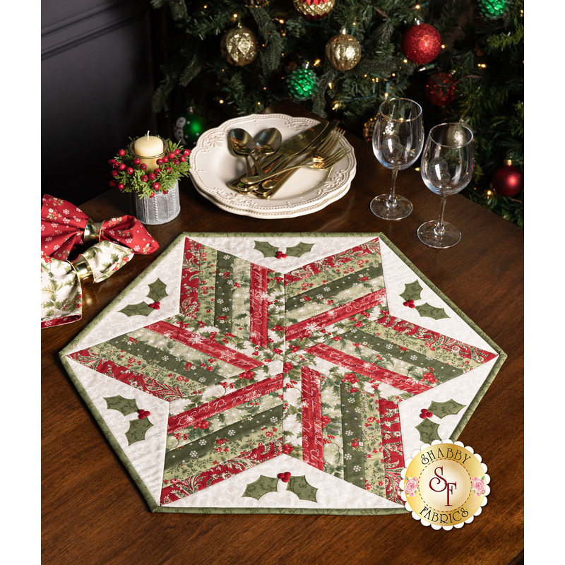 The completed 60 Degree Diamond Table Topper, colored in red, green, and white fabrics from the A Christmas Carol collection by Moda Fabrics, staged on a rustic wood table with coordinating decor. A colorful Christmas tree peeks into frame from above.