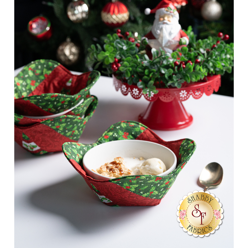 The completed bowl cozies, colored in red and green fabrics from the Yuletide Traditions collection, staged on a white table with coordinating holiday decor including a santa, garland, and Christmas tree.