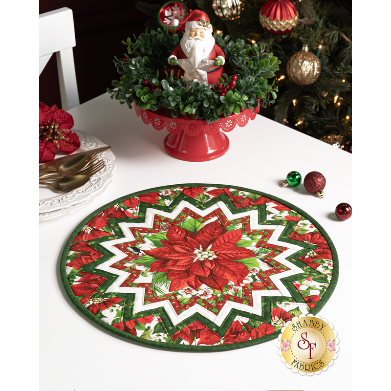 The completed table topper, colored in red and green fabrics from the Yuletide Traditions collection, staged on a white table with coordinating holiday decor including a santa, garland, and Christmas tree.