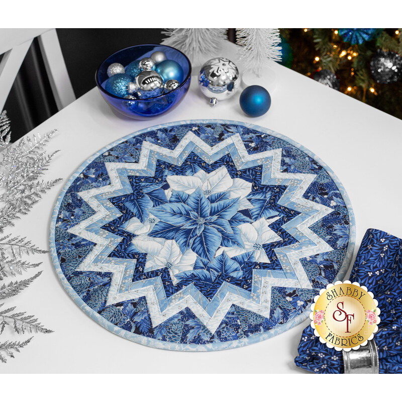 A shot of the completed Point of View Folded Star Table Topper Kit in fabrics from the Holiday Flourish - Festive Finery collection, a series of wintry blue metallic prints. It is staged on a white table with coordinating blue and winter decor.