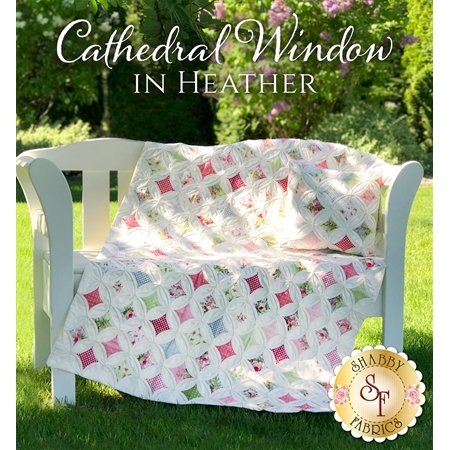 White and pastel pink Cathedral Window quilt draped over a white bench in a grassy park.
