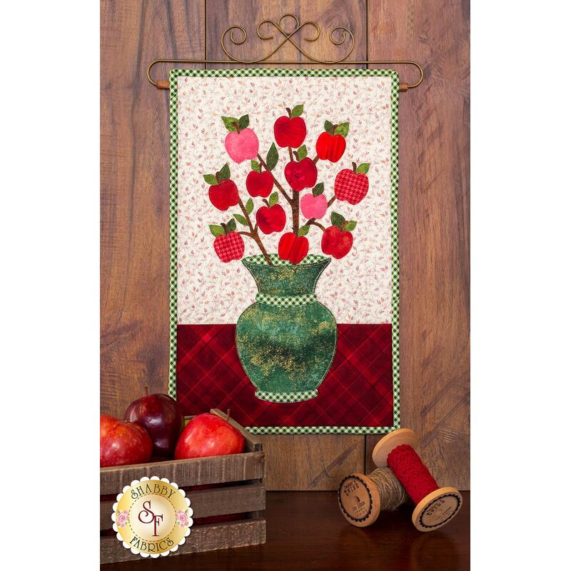 Mottled green vase filled with branches on which tiny red apples are hanging.