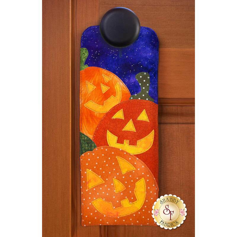 Door hanger kit for A-door-naments October with three smiling jack-o-lanterns on violet fabric.