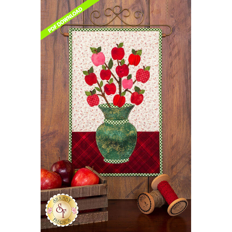 Mottled green vase filled with branches on which tiny red apples are hanging.