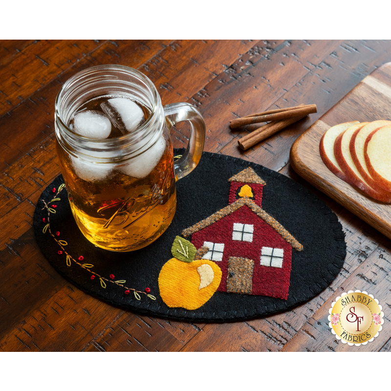 Small black wool mat with a school house and yellow apple stitched down, topped with a glass of tea